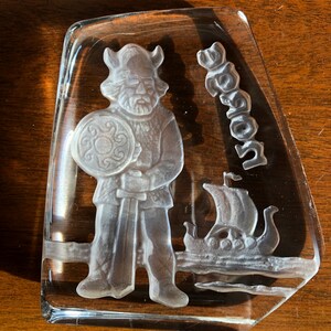 May include: A clear glass paperweight with a frosted design of a Viking warrior holding a sword and shield, with a Viking ship in the background. The word "Viking" is etched into the glass.
