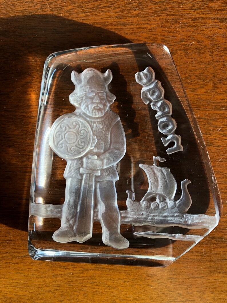 May include: A clear glass paperweight with a frosted design of a Viking warrior holding a sword and shield, with a Viking ship in the background. The word "Viking" is etched into the glass.