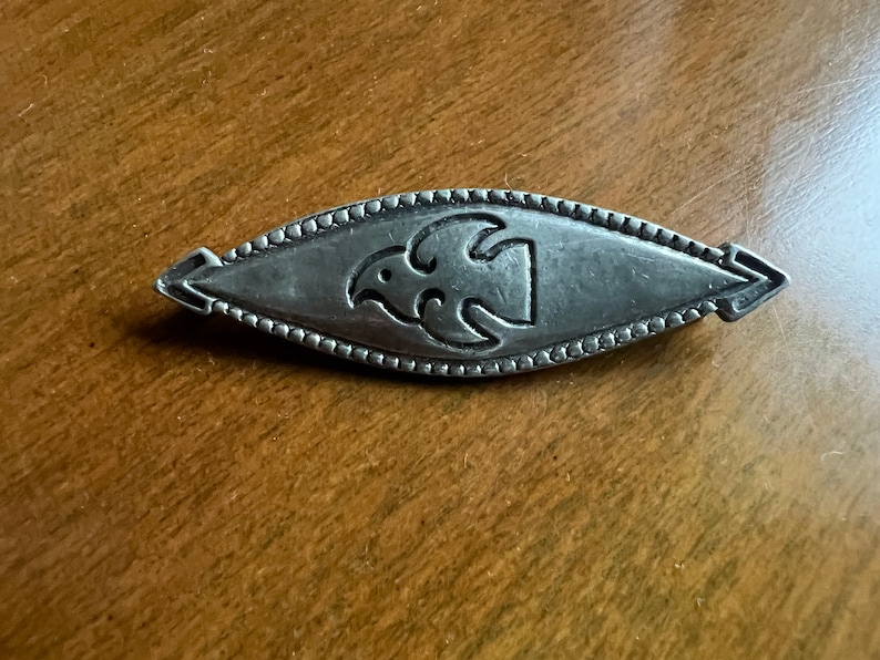 May include: A silver brooch with a stylized bird design. The brooch is oval shaped and has a beaded border.