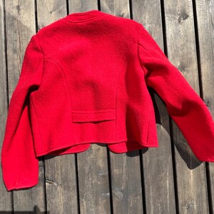 Puede incluir: Una chaqueta de lana roja de corte corto y mangas largas. La chaqueta tiene un cierre de bot&oacute;n &uacute;nico en el cuello y un bolsillo en la espalda.