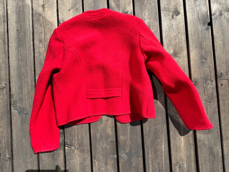 Puede incluir: Una chaqueta de lana roja de corte corto y mangas largas. La chaqueta tiene un cierre de bot&oacute;n &uacute;nico en el cuello y un bolsillo en la espalda.