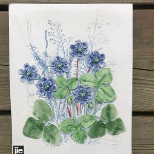 May include: A white ceramic tile featuring a blue and green floral design. The tile depicts a cluster of blue flowers with green leaves and stems. The tile is signed "Carl" in the bottom right corner and bears a "j.i.e. Sandtofta Sweden" logo in the bottom left corner.