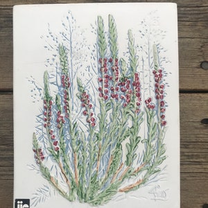 May include: A white ceramic tile featuring an embossed design of a group of green and red heather plants. The tile is signed "A. S" in the bottom right corner and has a "JIE" logo in the bottom left corner.