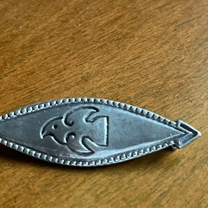 May include: A silver brooch with a stylized bird design. The brooch is oval shaped and has a beaded edge.