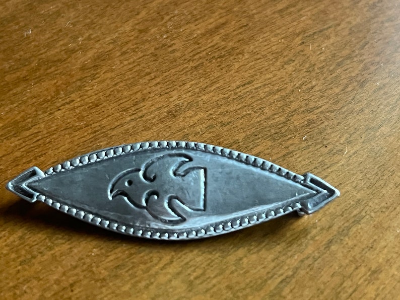 May include: A silver brooch with a stylized bird design. The brooch is oval shaped and has a beaded edge.