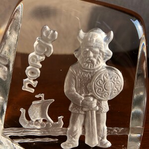 May include: A clear glass paperweight with a white etched design of a Viking warrior holding a sword and shield. The word "Norge" is etched in the background.