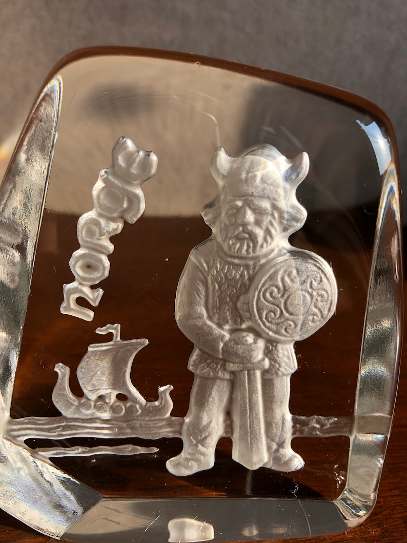 May include: A clear glass paperweight with a white etched design of a Viking warrior holding a sword and shield. The word "Norge" is etched in the background.