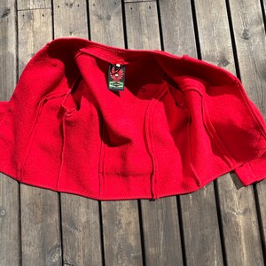 Puede incluir: Chaqueta de lana roja con una etiqueta cosida que dice "The Eagle's Nest". La chaqueta est&aacute; desabrochada y colocada sobre una superficie de madera.