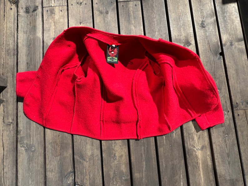 Puede incluir: Chaqueta de lana roja con una etiqueta cosida que dice "The Eagle's Nest". La chaqueta est&aacute; desabrochada y colocada sobre una superficie de madera.