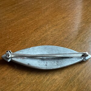May include: A silver pewter brooch in the shape of a leaf with a pin on the back. The brooch is engraved with the words "Vinje Pewter Made in Norway".