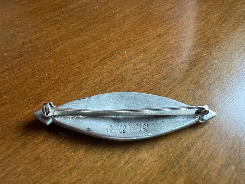 May include: A silver pewter brooch in the shape of a leaf with a pin on the back. The brooch is engraved with the words "Vinje Pewter Made in Norway".