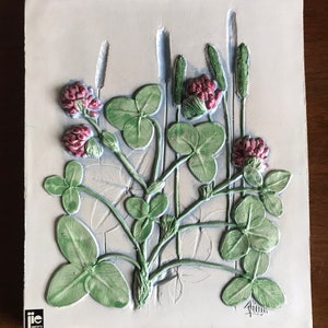 May include: A rectangular, off-white ceramic tile with a raised relief of clover and other plants. The clover has green leaves and pink flower heads. The tile has a light blue border and is signed in the bottom right corner.