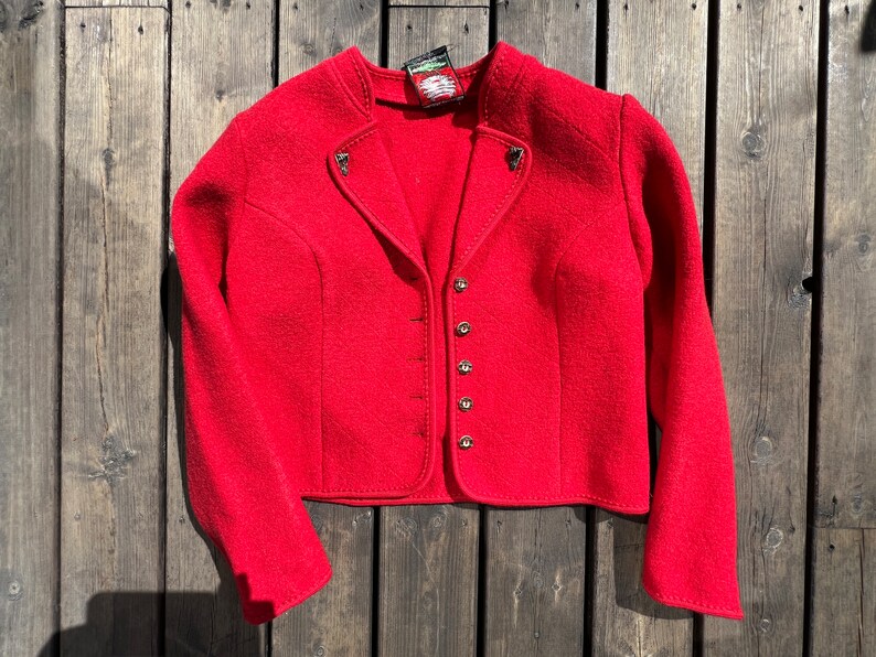 Puede incluir: Una chaqueta de lana roja con cierre de botones y una peque&ntilde;a etiqueta en el cuello. La chaqueta tiene un estilo tradicional con una longitud ligeramente recortada.