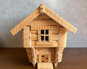 May include: A small wooden model of a house with a gabled roof, a single window, and a door. The house is made of logs and has a rustic appearance.