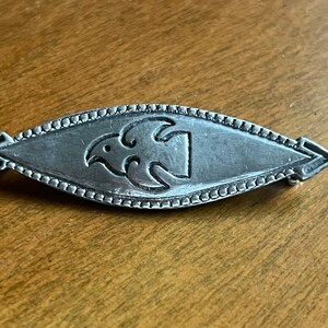 May include: A silver brooch with a stylized bird design in the center. The brooch is oval shaped and has a beaded border.