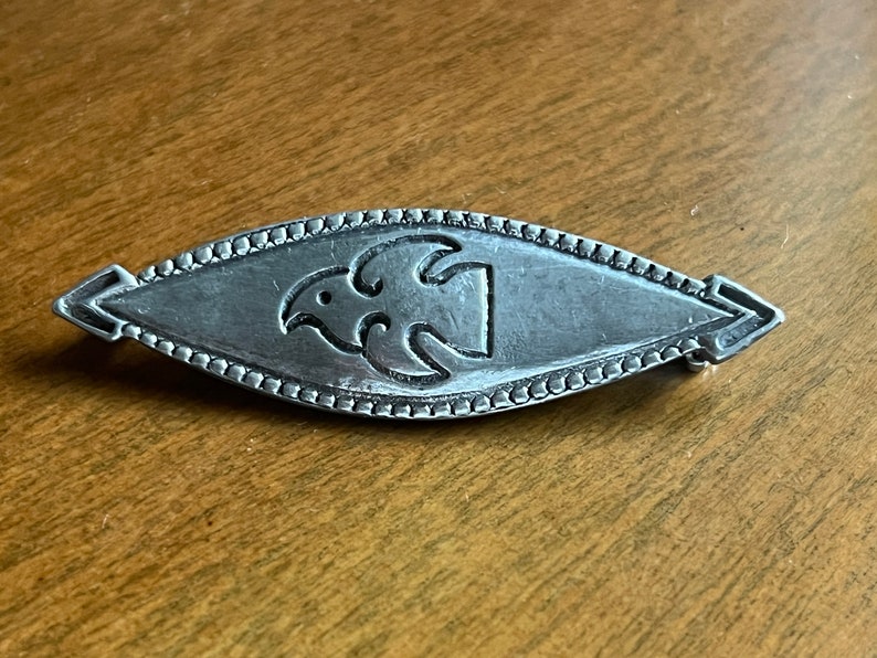 May include: A silver brooch with a stylized bird design in the center. The brooch is oval shaped and has a beaded border.