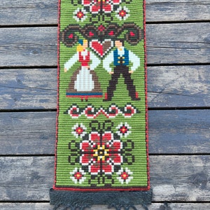 May include: A green, red, and white embroidered wall hanging with a folk art design featuring a couple in traditional clothing. The hanging is attached to a black metal hook.