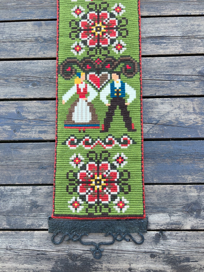 May include: A green, red, and white embroidered wall hanging with a folk art design featuring a couple in traditional clothing. The hanging is attached to a black metal hook.