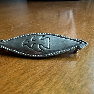 May include: A silver brooch with a stylized bird design in the center. The brooch is oval shaped and has a beaded edge.