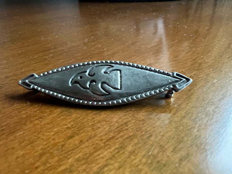 May include: A silver brooch with a stylized bird design in the center. The brooch is oval shaped and has a beaded edge.