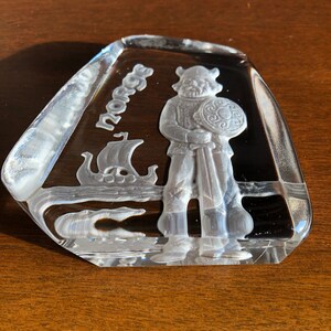 May include: A clear glass paperweight with a raised design of a Viking warrior holding a sword and shield. The paperweight is inscribed with the word "Norge".