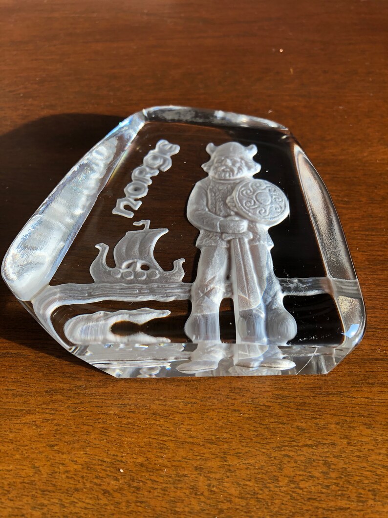 May include: A clear glass paperweight with a raised design of a Viking warrior holding a sword and shield. The paperweight is inscribed with the word "Norge".