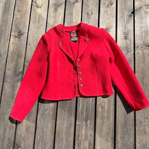 Puede incluir: Una chaqueta de lana roja con cuello alto, cierre de botones y botones decorativos en los hombros. La chaqueta tiene una longitud recortada y mangas largas.