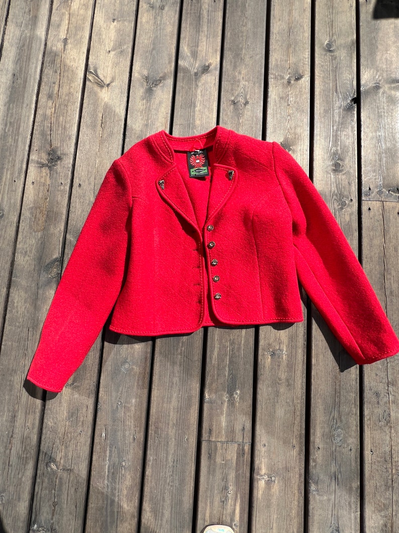 Puede incluir: Una chaqueta de lana roja con cuello alto, cierre de botones y botones decorativos en los hombros. La chaqueta tiene una longitud recortada y mangas largas.