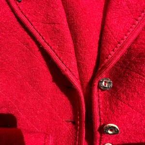 Puede incluir: Primer plano de una chaqueta roja con cremallera y botones. La chaqueta tiene una tela texturizada y un ribete negro. La etiqueta de la chaqueta dice "AUS &Ouml;STERREICH".
