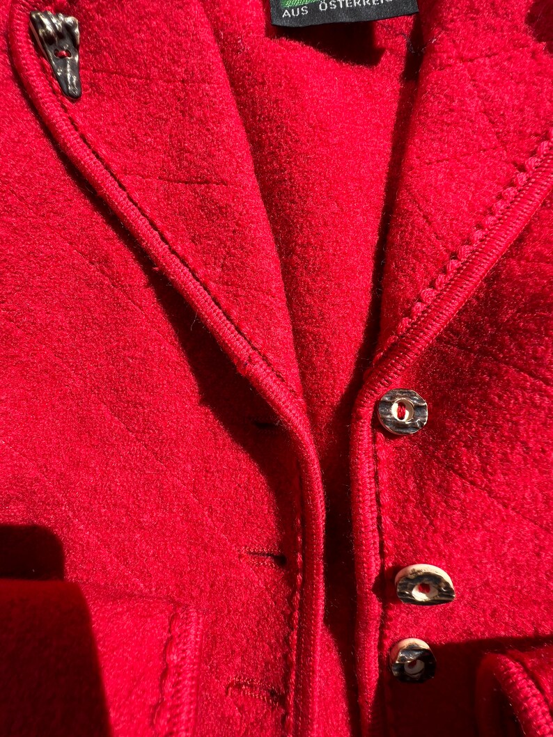 Puede incluir: Primer plano de una chaqueta roja con cremallera y botones. La chaqueta tiene una tela texturizada y un ribete negro. La etiqueta de la chaqueta dice "AUS &Ouml;STERREICH".