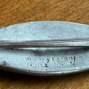 May include: A silver-colored pewter brooch in the shape of a fish. The brooch has a pin on the back and the words "Yinn-Der Made in Pewter Norway" are engraved on the bottom.
