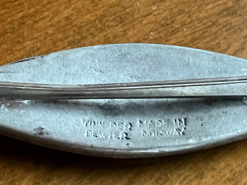 May include: A silver-colored pewter brooch in the shape of a fish. The brooch has a pin on the back and the words "Yinn-Der Made in Pewter Norway" are engraved on the bottom.