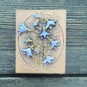 May include: A square, light brown ceramic tile with a relief of bluebell flowers. The flowers are light blue with gold stems, set within a circular border. The tile is against a weathered wood background.