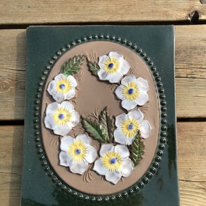 May include: A ceramic tile with a green border and a brown centre. The centre features a raised design of white flowers with yellow centres and green leaves.