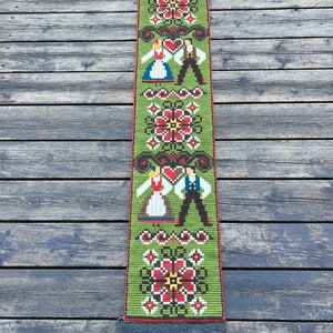 May include: A green tapestry with a folk art design featuring two figures in traditional clothing, flowers, and hearts. The tapestry is hanging from a black metal rod.