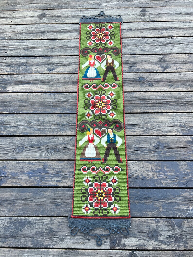 May include: A green tapestry with a folk art design featuring two figures in traditional clothing, flowers, and hearts. The tapestry is hanging from a black metal rod.