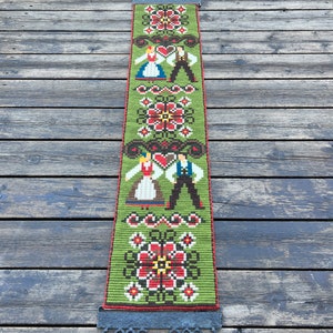 May include: A green tapestry with a traditional folk design featuring two figures in traditional clothing and floral patterns. The tapestry is hanging from a black metal rod.