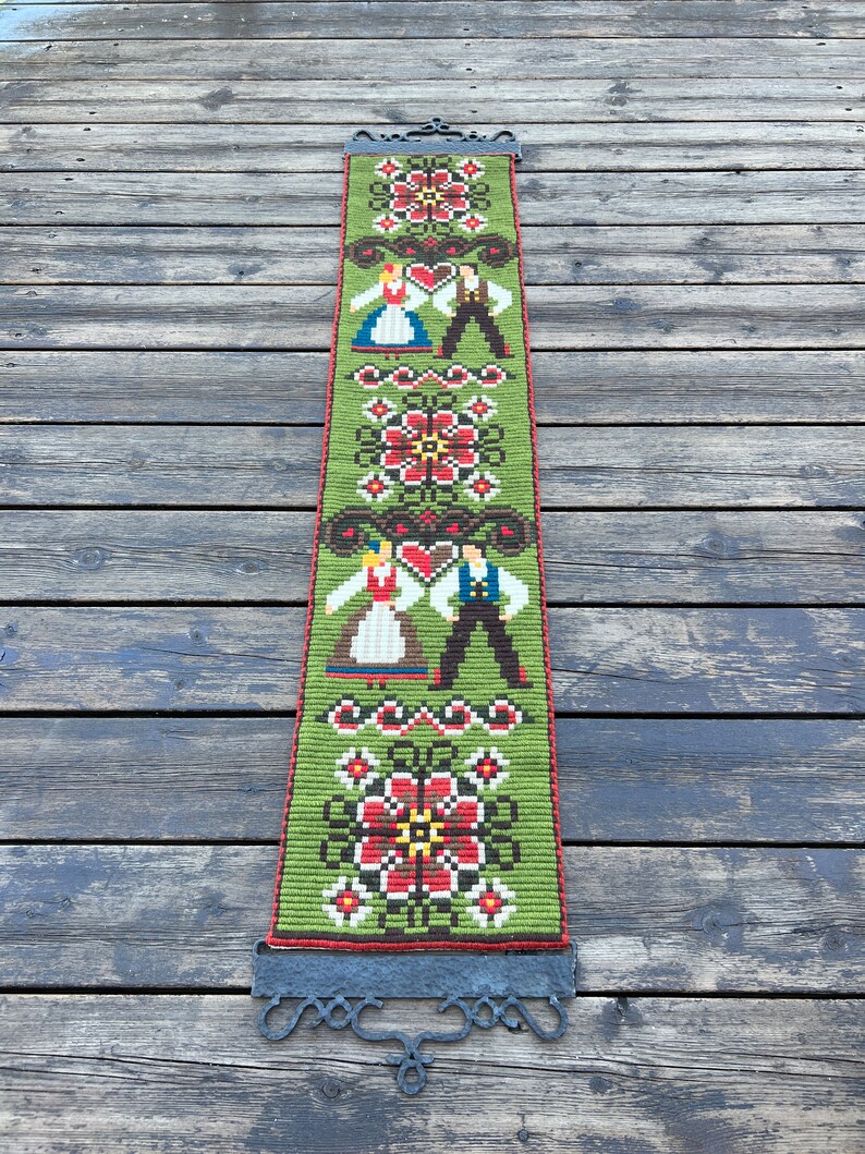 May include: A green tapestry with a traditional folk design featuring two figures in traditional clothing and floral patterns. The tapestry is hanging from a black metal rod.