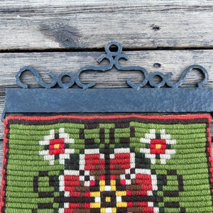 May include: A green, brown, red, and white tapestry with a floral design hangs from a black metal frame. The frame has a decorative scroll design at the top.