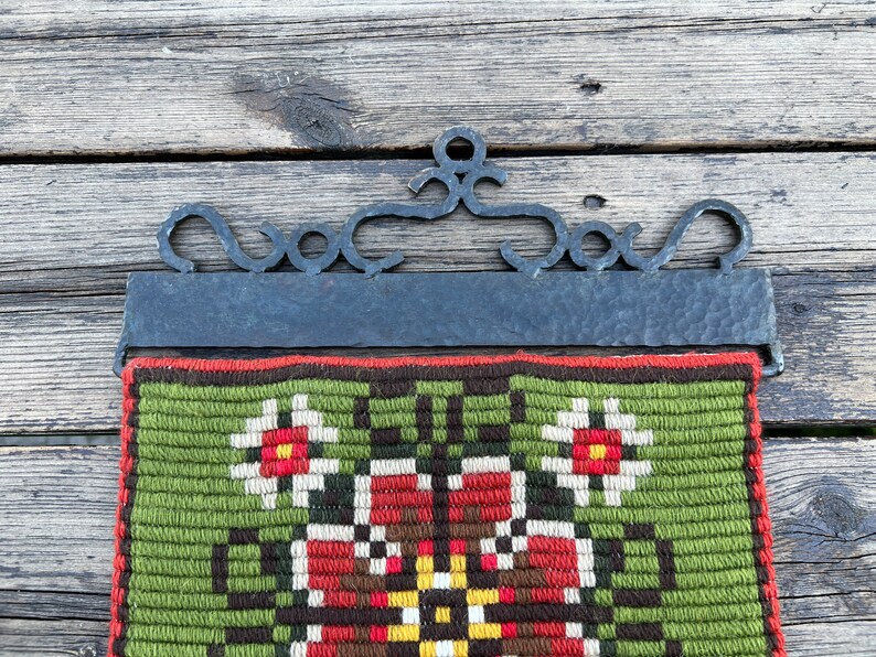 May include: A green, brown, red, and white tapestry with a floral design hangs from a black metal frame. The frame has a decorative scroll design at the top.