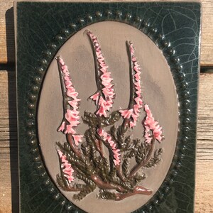 May include: A ceramic tile with a green border and a brown oval in the centre. The oval features a raised design of pink flowers and green leaves.