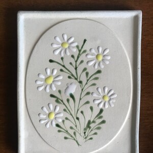 May include: A white ceramic tile featuring a raised design of white daisies with yellow centres and green stems. The tile is framed by a white border.