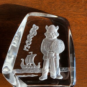 May include: A clear glass paperweight with a raised design of a Viking warrior holding a sword and shield. The word "Norse" is etched into the glass.