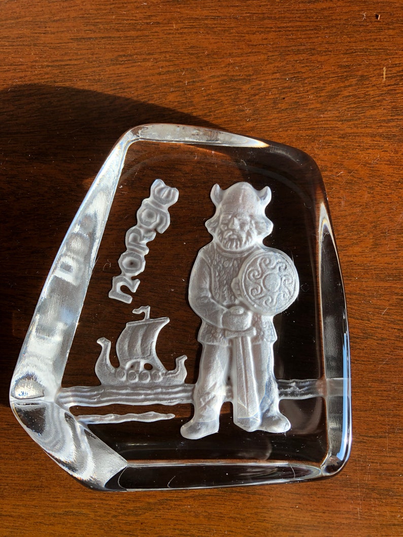 May include: A clear glass paperweight with a raised design of a Viking warrior holding a sword and shield. The word "Norse" is etched into the glass.