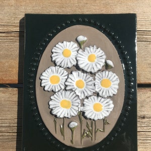 May include: A ceramic tile with a dark green border and a brown oval in the centre. The oval features a cluster of white daisies with yellow centres, arranged in a bouquet.