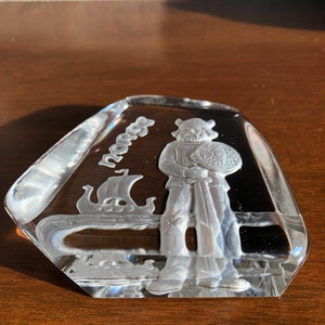 May include: A clear glass paperweight with an etched design of a Viking warrior holding a sword and shield. The word "Norge" is etched on the paperweight.