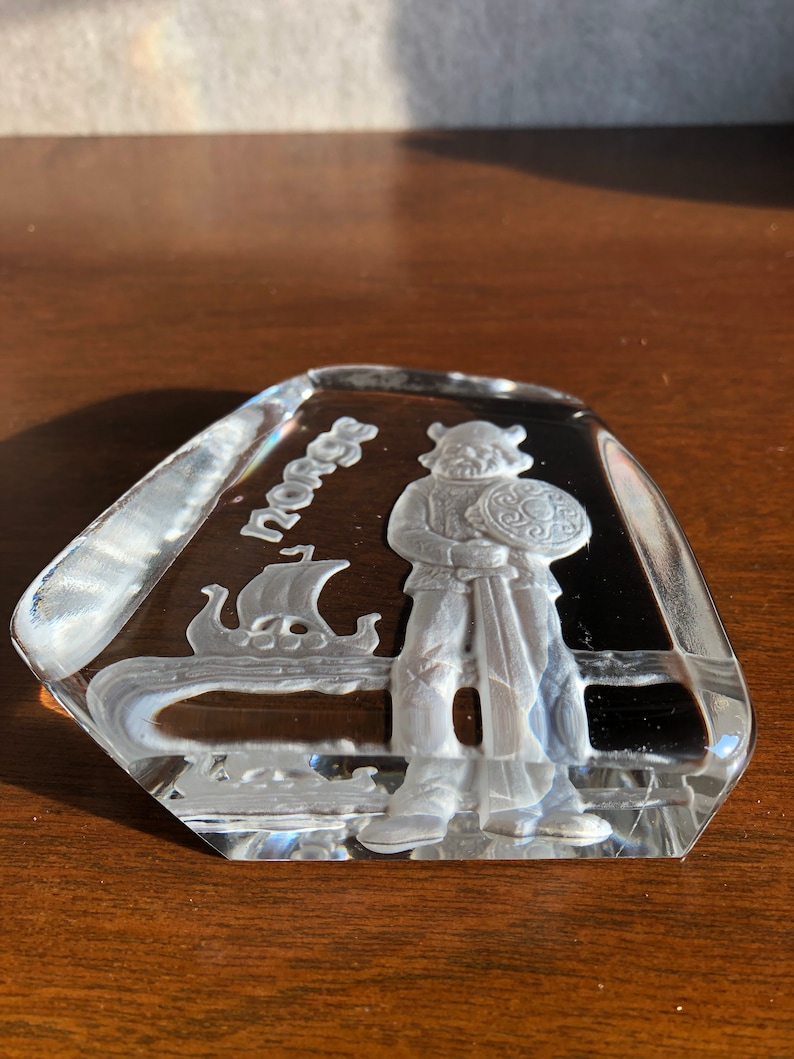 May include: A clear glass paperweight with an etched design of a Viking warrior holding a sword and shield. The word "Norge" is etched on the paperweight.