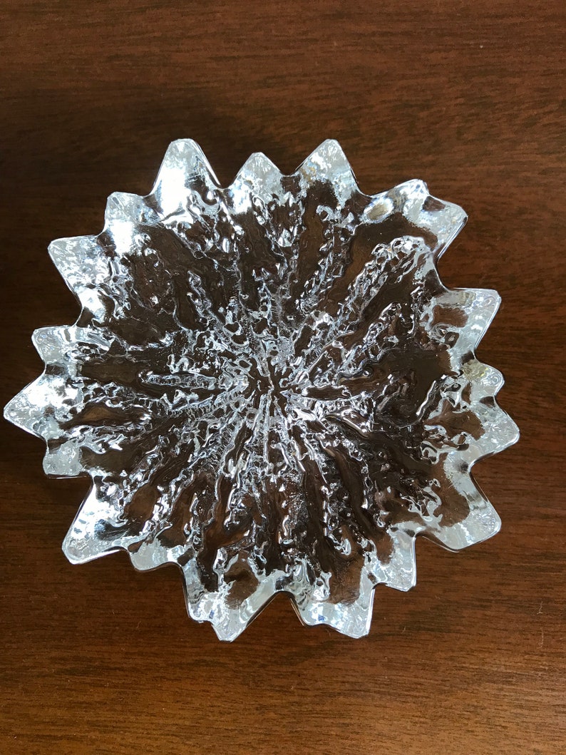 Pu&ograve; includere: Un piatto di vetro trasparente con un design a stella. Il piatto ha una superficie testurizzata e un bordo smerlato.