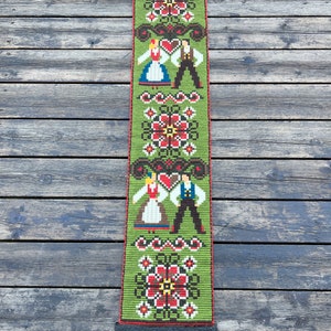 May include: A green tapestry with a folk art design featuring two figures in traditional clothing, red and white flowers, and a black metal hanging rod.