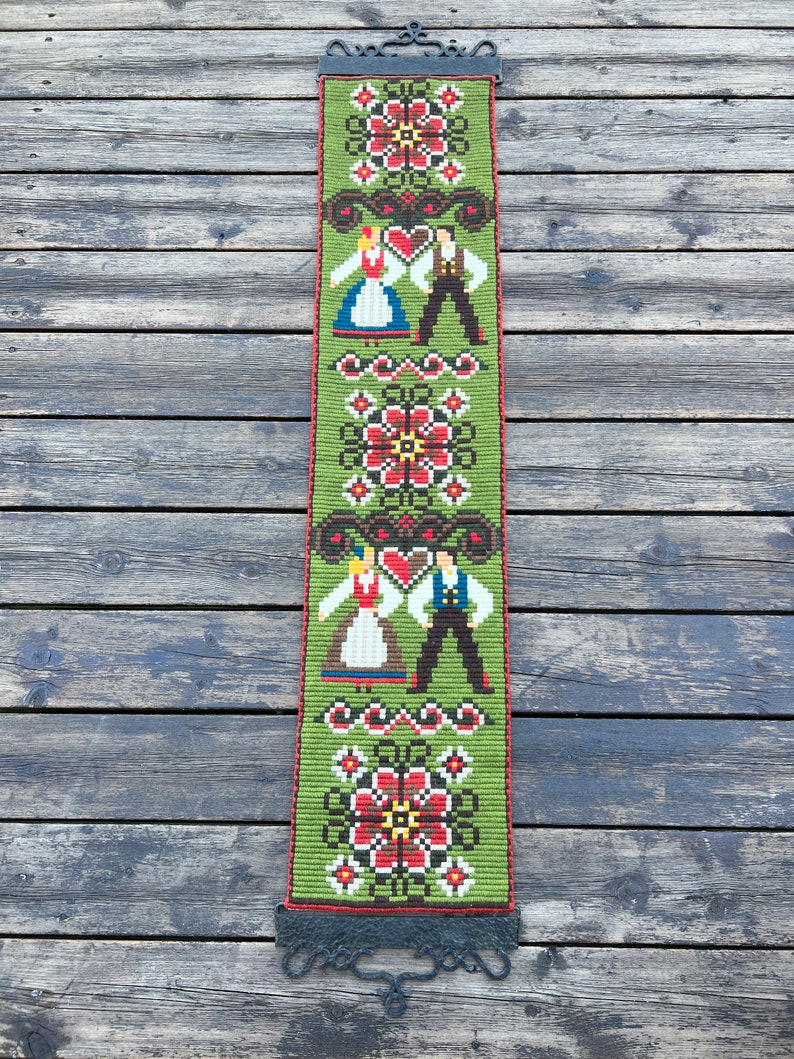 May include: A green tapestry with a folk art design featuring two figures in traditional clothing, red and white flowers, and a black metal hanging rod.